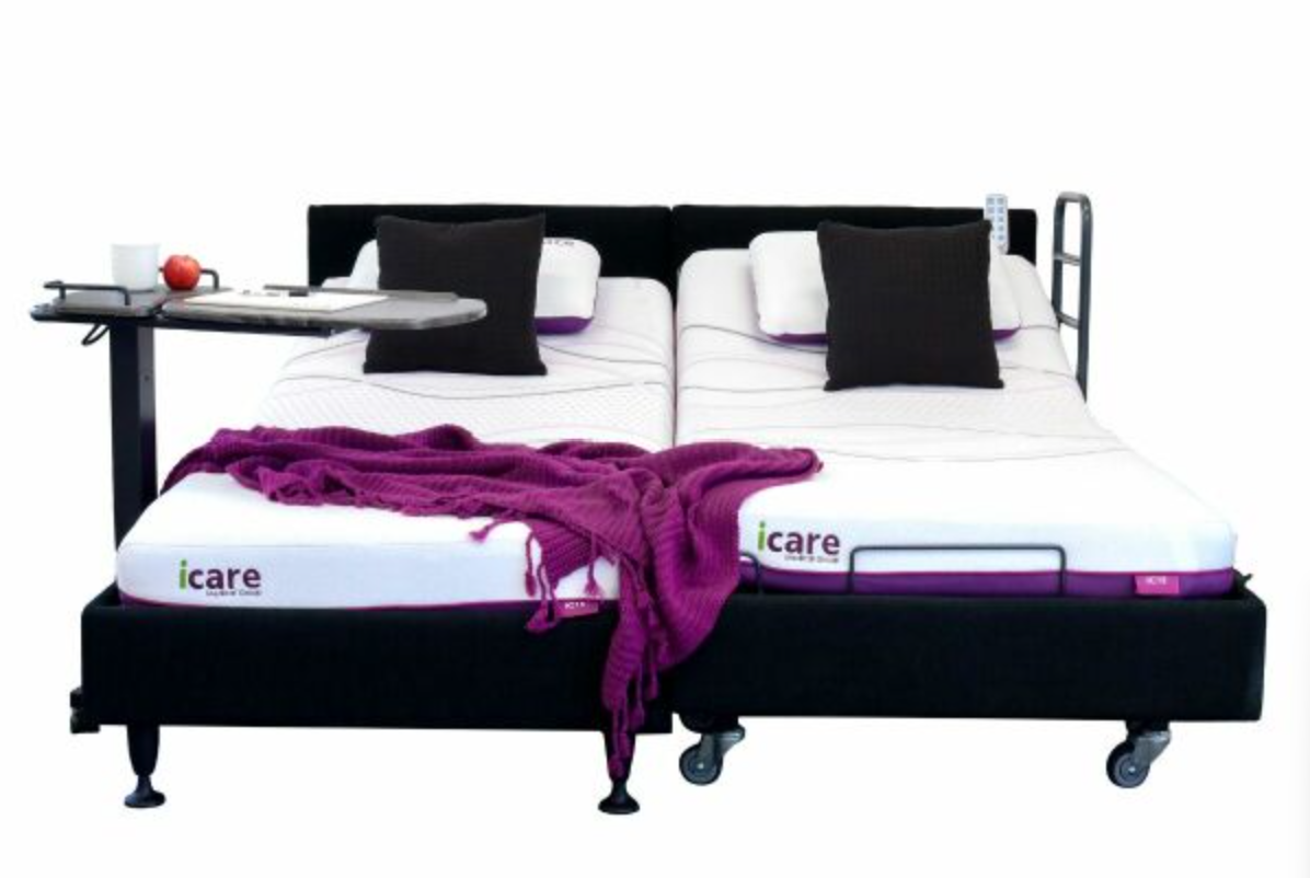 An adjustable dual-twin electric bed frame with a black upholstered base, white mattresses, black pillows, a purple throw blanket, and an attached side table on a white background.